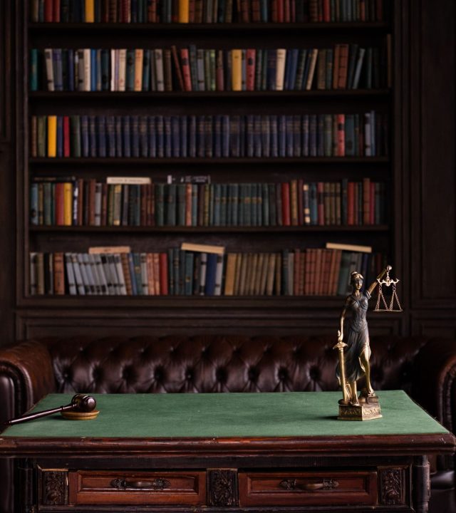 A classic study room with shelves of books, a gavel, and Lady Justice figurine on a green table.